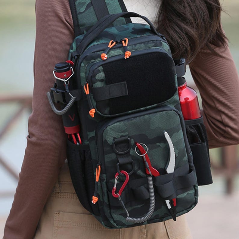 May include: A camouflage-patterned sling bag with multiple pockets and attachments. The bag features a black main compartment, orange accents, and a red water bottle in a side pocket. Additional items include a knife and a red carabiner.