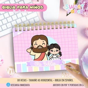 Może przedstawiać: Różowy notatnik w spiralnej oprawie z ilustracjami Jezusa i dziewczynki. Złote pierścienie i hiszpański tekst: "Mi Biblia" i "Biblia Para Niños". Format A5.