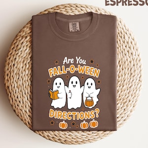 May include: Espresso-colored t-shirt with the text "Are You Fall-O-Ween Directions?" and three cartoon ghosts. One ghost holds a book, another a thumbs-up, and the third a pumpkin-shaped treat bag. Orange pumpkins and stars complete the design.