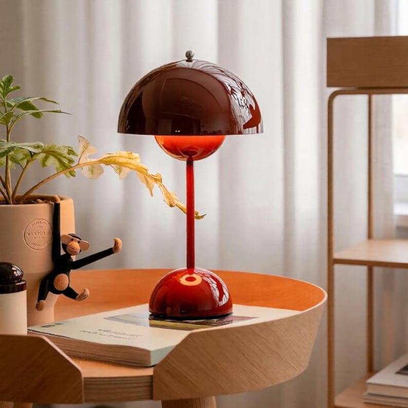 K&ouml;nnte beinhalten: Eine moderne, rote Tischlampe mit einem kuppelf&ouml;rmigen Schirm. Die Lampe hat einen runden Sockel und einen schlanken Stiel. Der Schirm ist dunkelrotbraun und der Sockel ist heller rot. Die Lampe steht auf einem Holztisch.