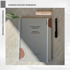 May include: A spiral-bound workbook titled "Business Strategy Workbook" with the text "Branding & Positionierung." A black pen and a pink highlighter sit next to the workbook. The cover is light gray with brown and white accents.