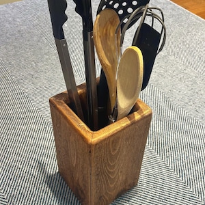 May include: A wooden utensil holder containing various kitchen tools. The holder is a square, stained wooden box. The tools include a black slotted spoon, a whisk, tongs, and wooden spoons. The holder sits on a grey and white striped surface.