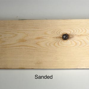 K&ouml;nnte beinhalten: Ein helles, geschliffenes Holzbrett mit einem markanten Knoten in der Mitte. Die Holzmaserung ist sichtbar und die Oberfl&auml;che ist glatt. Das Wort "Sanded" ist unten im Bild schwarz gedruckt.