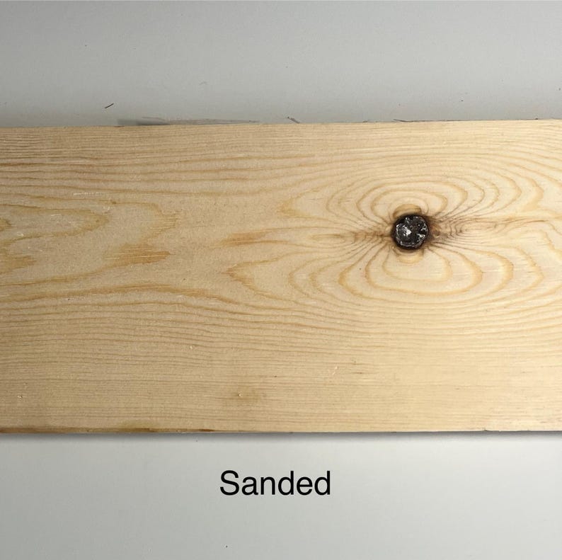 K&ouml;nnte beinhalten: Ein helles, geschliffenes Holzbrett mit einem markanten Knoten in der Mitte. Die Holzmaserung ist sichtbar und die Oberfl&auml;che ist glatt. Das Wort "Sanded" ist unten im Bild schwarz gedruckt.