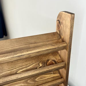 K&ouml;nnte beinhalten: Nahaufnahme eines Holzregals mit vier horizontalen Regalb&ouml;den. Das Holz hat eine warme, braune Beize, und die Maserung ist sichtbar. Das Regal ist mit einer einfachen, rustikalen &Auml;sthetik gestaltet, perfekt zum Pr&auml;sentieren von Gegenst&auml;nden.