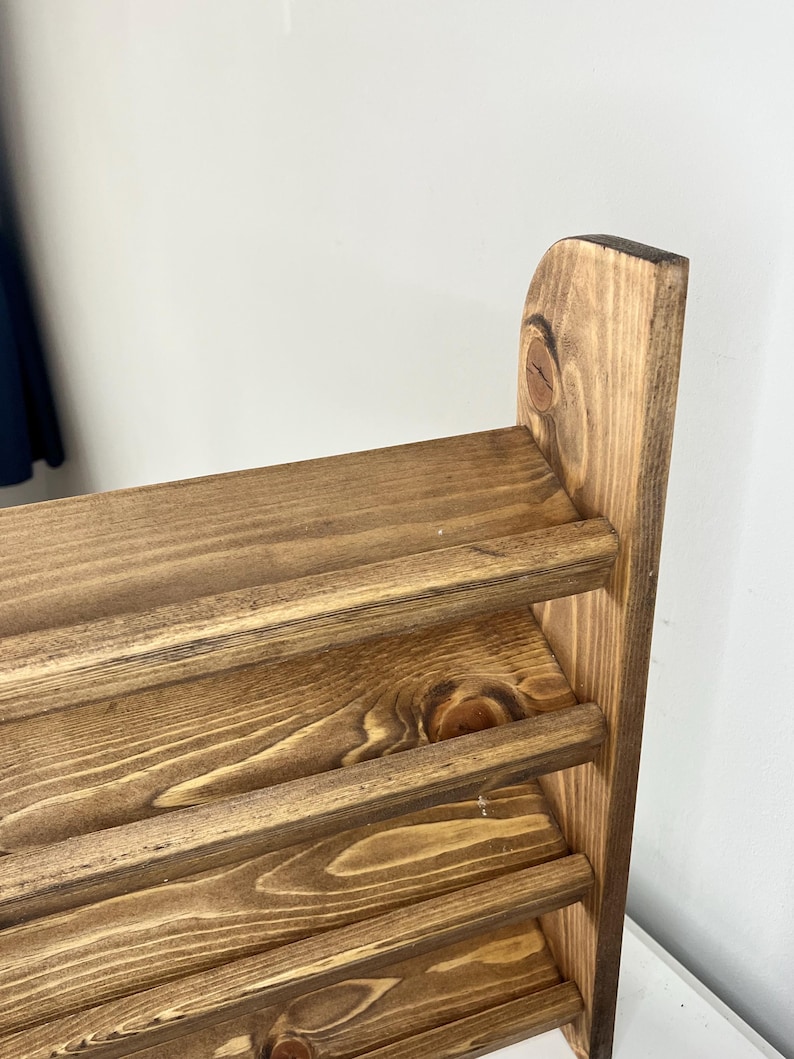 K&ouml;nnte beinhalten: Nahaufnahme eines Holzregals mit vier horizontalen Regalb&ouml;den. Das Holz hat eine warme, braune Beize, und die Maserung ist sichtbar. Das Regal ist mit einer einfachen, rustikalen &Auml;sthetik gestaltet, perfekt zum Pr&auml;sentieren von Gegenst&auml;nden.