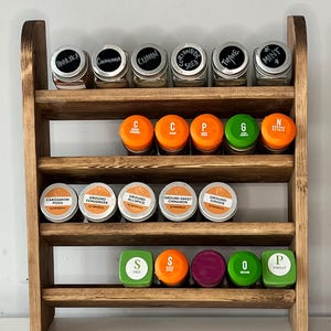 K&ouml;nnte beinhalten: Ein h&ouml;lzernes Gew&uuml;rzregal mit drei Regalen, das verschiedene Gew&uuml;rzgl&auml;ser und -beh&auml;lter enth&auml;lt. Das oberste Regal zeigt klare Gl&auml;ser mit schwarzen Tafellabels, darunter "Paprika" und "Cinnamon". Das mittlere Regal hat orangefarbene Deckel.