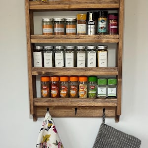 May include: A wooden spice rack with multiple spice jars and bottles of oil and sauces. The rack has three shelves and hooks below. A floral tea towel and a grey oven glove hang from the bottom.