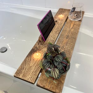 May include: A wooden bath tray with a slot for a tablet, a small potted plant, two lit candles, and a glass. The tray is made of reclaimed wood and spans the width of a white bathtub. The glass has text on it.