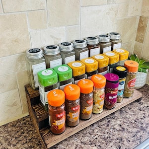 May include: A three-tiered wooden spice rack displaying numerous spice jars with different coloured lids and labels. The jars are neatly arranged, showcasing a variety of herbs and spices. The rack is on a countertop with a neutral-toned backsplash.