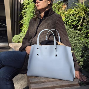 May include: A light blue tote bag with two top handles, silver-toned metal rivets at the base of the handles, and the letters EZ embossed in the centre. The bag has a structured, open-top design with visible stitching along the side edges.