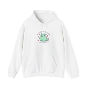 May include: White hoodie with a front pocket and drawstring hood. The front features a cartoon cat illustration with the text "I'M TRYING TO STAY CUTE DON'T TEST IT". The cat is green with pink accents and is holding a knife.
