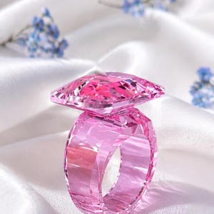 Può includere: Un anello rosa sfaccettato realizzato in un unico pezzo di materiale traslucido. L'anello presenta una fascia larga e spessa e una grande sezione superiore di forma quadrata con bordi geometrici netti che catturano la luce.