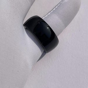 Può includere: Un anello nero lucido con un design liscio e arrotondato. L'anello è una fascia solida, che riflette la luce e appare elegante. Il gioiello è mostrato su una superficie bianca.
