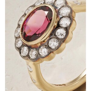 May include: A gold ring featuring a large oval, reddish-pink gemstone at the center, surrounded by a halo of small, clear gemstones. The ring's band is gold, with additional small gemstones set into the band.