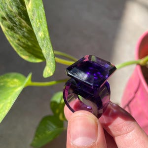 May include: A deep purple ring featuring a large, faceted, rectangular-cut gemstone set atop a thick, matching purple band. The ring is crafted from a translucent, glass-like material that catches the light, creating sharp, geometric reflections.