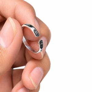 Könnte beinhalten: Ein silberfarbener Ring mit den Buchstaben "M" und "J" in Schwarz. Der Ring wird zwischen Fingern vor weißem Hintergrund gehalten. Der Ring hat ein schlichtes, offenes Design.