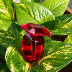 Op de afbeelding: Een felrode ring gemaakt uit één stuk doorschijnend, gefacetteerd materiaal. De ring heeft een groot, vierkant bovendeel met scherpe, geometrische randen en een brede, hoekige band die het licht opvangt en dieprode reflecties creëert.