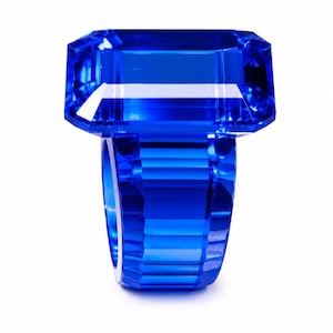 Puede incluir: Un anillo de color azul vibrante con una parte superior grande, rectangular y facetada, similar a una piedra preciosa, sobre una banda ancha y multifacetada. La pieza está hecha de un material azul profundo, translúcido y con bordes afilados.