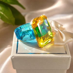 May include: A chunky ring featuring a translucent blue band and a large, rectangular, faceted gemstone that transitions from yellow to green. The ring is positioned on top of a square white gift box adorned with a small, matching white fabric bow.