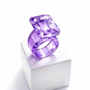 Könnte beinhalten: Ein großer, transparenter, violetter Ring mit einem rechteckigen Edelstein. Der Ring wird auf einer weißen quadratischen Box vor weißem Hintergrund präsentiert. Der Edelstein hat einen leuchtenden Lilaton, und der Ring ist dick und glatt.