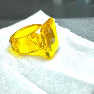 Op de afbeelding: Een grote, gele ring met een facetgeslepen, rechthoekige edelsteen. De ring is gemaakt van een transparant materiaal en ligt op een witte, getextureerde ondergrond. De band van de ring heeft een geometrisch ontwerp.