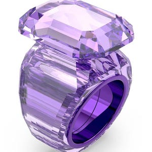 Może przedstawiać: Fasetowany, fioletowy kryształowy pierścionek z dużym, prostokątnym kamieniem wierzchnim. Opaska pierścionka i kamień wierzchni mają pasujący odcień lawendy, tworząc spójny i elegancki design. Pierścionek jest na białym tle.