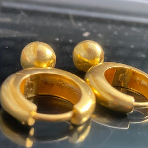 Könnte beinhalten: Nahaufnahme von zwei Paar goldfarbenen Ohrringen. Ein Paar ist hufeisenförmig, das andere sind kleine, runde Kugelstecker. Die Ohrringe werden auf einer reflektierenden Oberfläche präsentiert.
