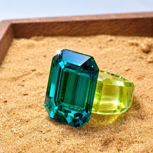 Op de afbeelding: Een ring met een grote, rechthoekige, smaragdgeslepen blauwgroene edelsteen op een doorschijnende, felgele band. Het sieraad heeft een hoogglans afwerking met scherpe, gefacetteerde randen die het licht vangen, wat zorgt voor een contrast tussen de blauwgroene steen en de gele ringbasis.