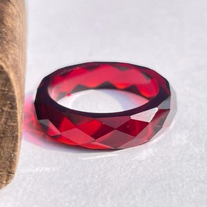 Peut inclure: Une bague rouge foncé à facettes. La surface de la bague est taillée avec de nombreuses facettes géométriques, créant un effet scintillant. La bague est une bande solide de matériau rouge, avec un intérieur poli. C'est un bijou.