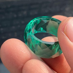 Puede incluir: Un anillo facetado de color verde esmeralda. El anillo se sostiene entre los dedos, mostrando su corte geométrico y su calidad transparente. El diseño del anillo presenta múltiples facetas, creando un efecto brillante. Es un accesorio vibrante.