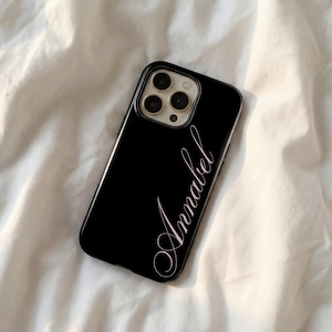 May include: A black phone case with the name "Annabel" in a cursive, light pink font. The case has a glossy finish and a camera cutout. The phone case is lying on a white, textured surface.