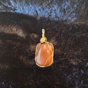 May include: A pendant featuring a reddish-brown stone wrapped in gold-colored wire. The stone has a smooth, polished appearance and is rectangular with rounded edges. The wire is intricately woven around the stone, creating a decorative frame and a loop for a chain.