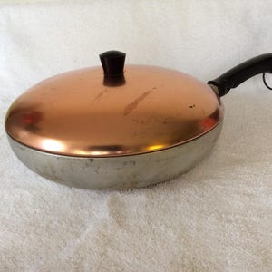 May include: A vintage copper-colored frying pan with a lid. The pan has a silver-colored base and a black handle. The lid has a black knob. The pan is sitting on a white towel.