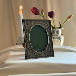 May include: A rectangular picture frame with an oval center opening and an ornate, embossed leaf pattern in a dark metallic finish. The frame features a deep green insert and stands upright on a smooth, draped white fabric surface.