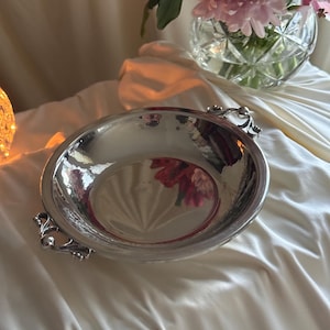 May include: A round, polished silver-toned metal bowl with a smooth, reflective interior surface. The bowl features two ornate, decorative handles on opposite sides, each with a detailed, scrolled metal design that curves outward from the rim.