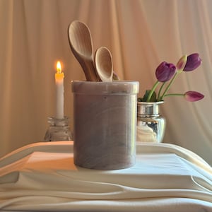 May include: A cylindrical light gray marble utensil holder with subtle veining, containing two light-colored wooden spoons of different sizes. The container has a smooth, polished finish and a thick, rounded rim at the top.