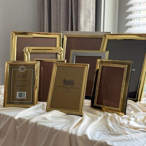 May include: A collection of rectangular photo frames in various sizes and finishes, including polished gold-tone metal, silver-tone metal, and textured brass. Several frames feature labels indicating sizes such as 4"x6" and 5"x7" with solid brass branding.