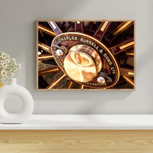 Peut inclure: Gros plan d'une impression encadrée d'une roue de machine à vapeur vintage. La roue est bordeaux foncé avec des accents dorés et un moyeu central en laiton. Le texte "CHARLES BURRELL & SONS LTD ENGINEERS ENGLAND" est visible.