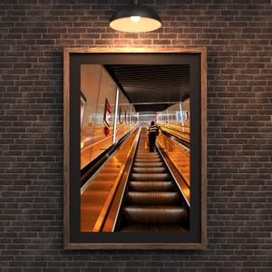 Può includere: Una stampa incorniciata di una persona su una scala mobile in una stazione della metropolitana. La scala mobile è di un colore dorato caldo e la persona indossa una camicia a righe. La stampa è su un muro di mattoni scuri.