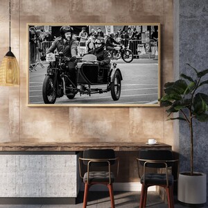 May include: Monochrome framed photograph of a classic motorcycle and sidecar racing. Two people in helmets and goggles are riding the motorcycle. The photograph is displayed on a wall above a wooden desk with two chairs, in a room.