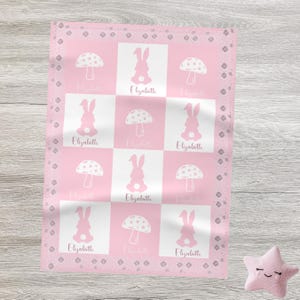 May include: A pink and white baby blanket with a repeating pattern of bunnies and mushrooms. Each square features a bunny or mushroom design, with the name "Elisabeth" printed below. The blanket has a decorative border and is next to a pink star-shaped pillow.