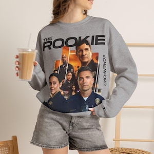 Könnte beinhalten: Ein graumeliertes Sweatshirt mit Rundhalsausschnitt und einem Grafikdruck der Fernsehserie The Rookie. Das Design enthält den Schriftzug The Rookie und #CHENFORD sowie eine Collage von Darstellern in Polizeiuniformen vor einem orangefarbenen Hintergrund.