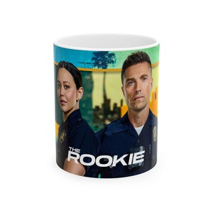 May include: A white ceramic mug featuring a graphic print of two police officers in dark blue uniforms. The text THE ROOKIE is printed in white at the bottom centre. The background shows a stylised city skyline with a palm tree and yellow accents.