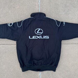 Pu&ograve; includere: Giacca nera con logo Lexus bianco sul retro e sulle maniche, con profili bianchi. La giacca ha un collo alto e un orlo a coste.