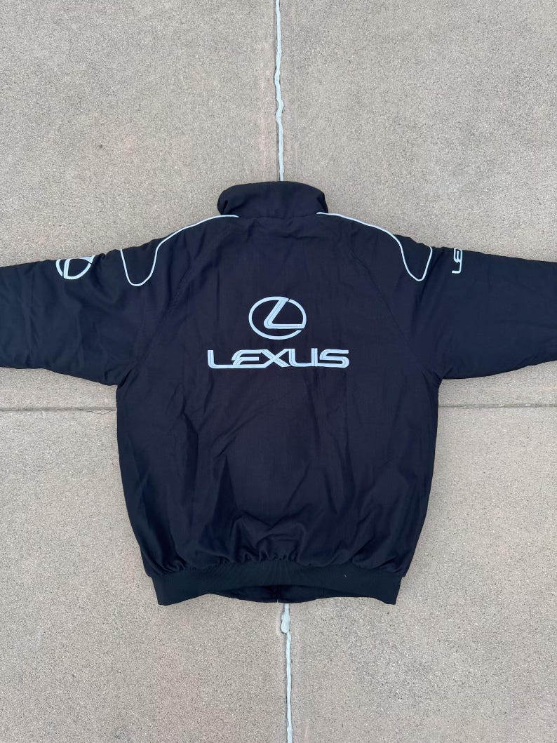 Pu&ograve; includere: Giacca nera con logo Lexus bianco sul retro e sulle maniche, con profili bianchi. La giacca ha un collo alto e un orlo a coste.