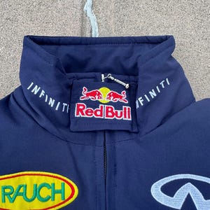 May include: Navy blue jacket with embroidered logos, including "Red Bull" and "Infiniti." The jacket features a high collar and a zipper closure. Additional logos include "RAUCH" and "GEOX."