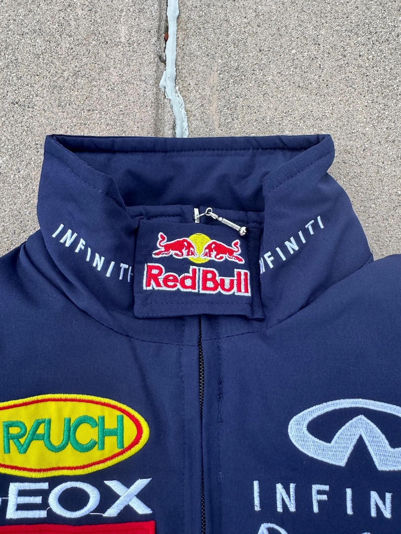 May include: Navy blue jacket with embroidered logos, including "Red Bull" and "Infiniti." The jacket features a high collar and a zipper closure. Additional logos include "RAUCH" and "GEOX."