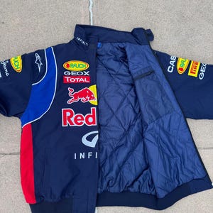 May include: Navy blue racing jacket with red and white accents. Features multiple sponsor logos, including "Red Bull" and "Infiniti." The jacket has a quilted interior and a zip-up front.