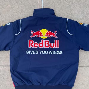 May include: Navy blue bomber jacket featuring the Red Bull logo in red and yellow. The jacket has white accents and the text "GIVES YOU WINGS" in white. The jacket is a sporty style.